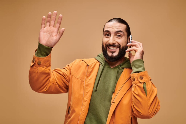 friendly middle eastern man with beard talking on smartphone on beige background, wave hand - Zdjęcie, obraz