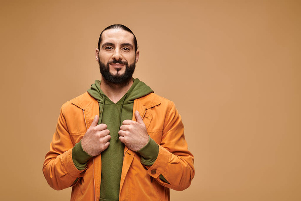 довольный ближневосточный мужчина с бородой, стоящий в повседневной одежде на бежевом фоне, глядя в камеру - Фото, изображение