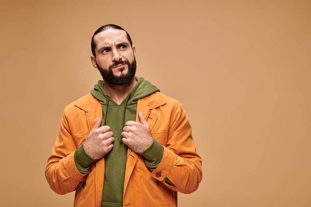 pensive middle eastern man with beard standing in casual attire on beige backdrop, looking away - Foto, afbeelding
