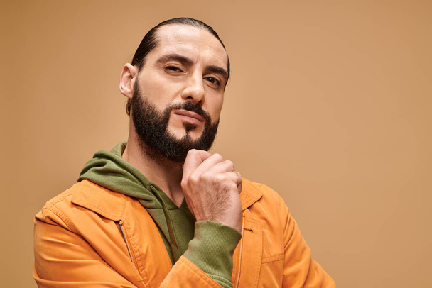 pensive middle eastern man touching beard and standing in casual attire on beige background - Photo, Image