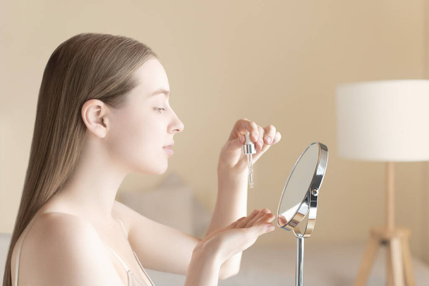 Mujer rubia caucásica aplica suero facial con un cuentagotas mientras se mira en el espejo. La mujer hidrata la piel, suaviza las arrugas, previene el acné, reduce la sensibilidad de la piel. Cuidado facial de la piel - Foto, imagen