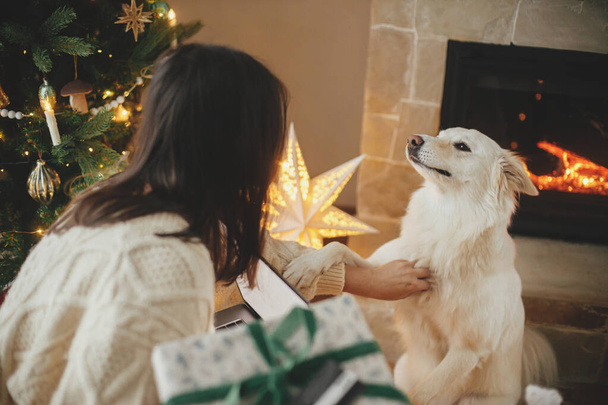 Mulher feliz segurando presentes de Natal elegantes com cartão de crédito e acariciando o cão branco bonito na sala de Natal decorada festiva com luzes. Férias de Natal e animal de estimação - Foto, Imagem