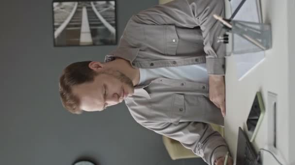 Genç beyaz adamın minimalist ofiste grafit duvarlar ve beyaz mobilyalarla çalışırken dizüstü bilgisayar kullanırken çekilmiş dikey belden yukarı pozu. - Video, Çekim