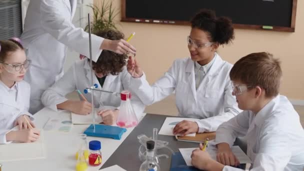 De arriba plano de diverso grupo de preadolescentes científicos en batas de laboratorio y su maestro haciendo experimentos químicos con líquidos en frasco de vidrio en el escritorio blanco en el aula moderna - Metraje, vídeo