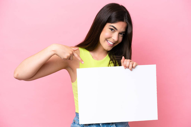 Young Brazilian woman isolated on pink background holding an empty placard with happy expression and pointing it - Valokuva, kuva