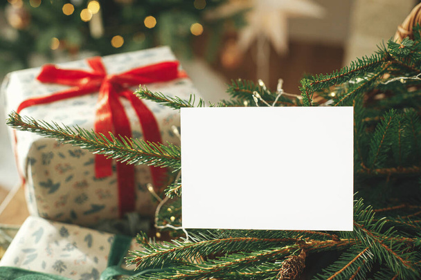 Χριστουγεννιάτικη κάρτα mock up. Άδειο ευχετήρια κάρτα στο παρασκήνιο των κομψών χριστουγεννιάτικα τυλιγμένα δώρα, κλαδιά ελάτης και εορταστικά χρυσά φώτα. Χώρος για κείμενο. Πρότυπο χαιρετισμού εποχής - Φωτογραφία, εικόνα