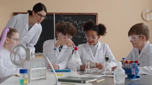 Gruppo di scolari preadolescenti diversi e la loro insegnante di chimica femminile tutti indossando camici bianchi fare esperimenti in classe di scienze moderne durante la lezione - Filmati, video