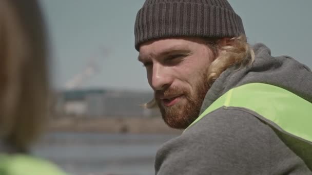 Über die Schulter eines jungen Umwelthelfers, der im Freien sitzt und mit einem Freund spricht, während er während der Aufräumarbeiten an der Küste eine Pause einlegt - Filmmaterial, Video
