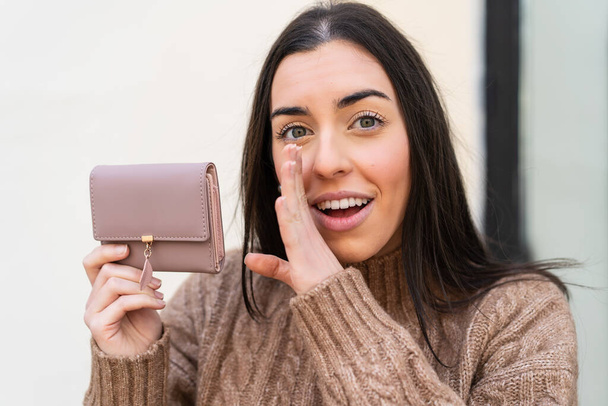 Mladá žena drží peněženku venku a něco si šeptá. - Fotografie, Obrázek