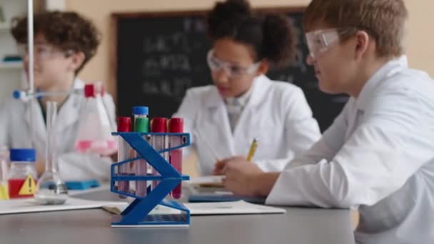 Pas up skupiny multiraciálních žáků v bílých laboratorních pláštích sedí za stolem s chemickým sklem na něm v moderní učebně mluví a studuje vědu - Záběry, video