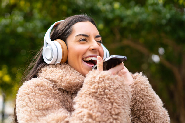Genç esmer kadın açık havada müzik dinliyor ve şarkı söylüyor. - Fotoğraf, Görsel