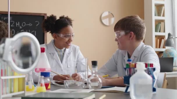 Очікування багаторасових дев'ятнадцяти однокласників в білих лабораторних халатах і захисних окулярах балакають під час проведення лабораторних експериментів під час уроку хімії в школі - Кадри, відео