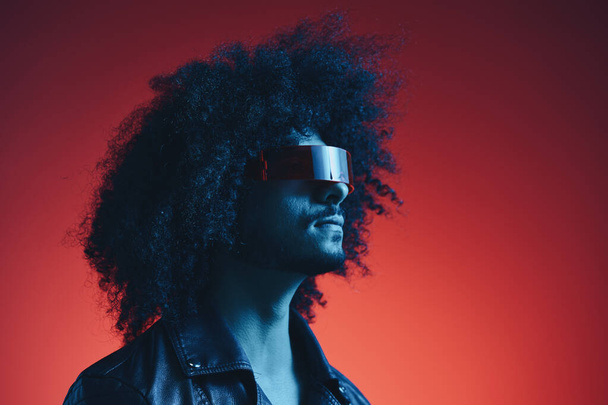 Portrait of fashion man with curly hair on red background with stylish glasses, multicultural, colored light, black leather jacket trend, modern concept. High quality photo - Photo, Image