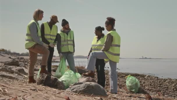 Colpo completo di gruppo di diversi attivisti ambientali in piedi con sacchi della spazzatura sulla costa e discutendo durante la pausa durante la raccolta della spazzatura - Filmati, video