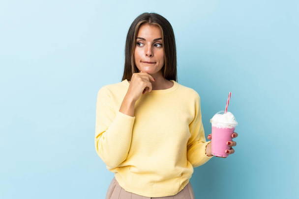 Jonge Uruguayaanse vrouw met aardbeien milkshake geïsoleerd op blauwe achtergrond met twijfels en denken - Foto, afbeelding