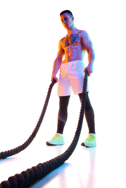 Muscular young man with fit, strong shirtless body standing with battle rope against white studio background in neon light. Concept of sport, active and healthy lifestyle, body care, fitness - Foto, Imagem