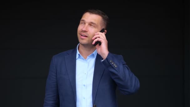 Close up portrait of handsome cheerful businessman speaking on mobile phone while standing outdoor near office center. Happy young confident man in suit chatting on smartphone. Real time concept. - Footage, Video