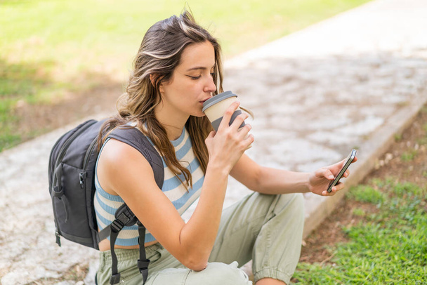 Młoda, ładna kobieta na świeżym powietrzu używa telefonu komórkowego i trzyma kawę na wynos - Zdjęcie, obraz