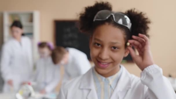 Zoom keskipitkällä lähikuva muotokuva iloinen Afrikkalainen Amerikan esikoulutyttö yllään valkoinen lab takki hymyillen kameran seisoo kirkas moderni koulu laboratorio - Materiaali, video