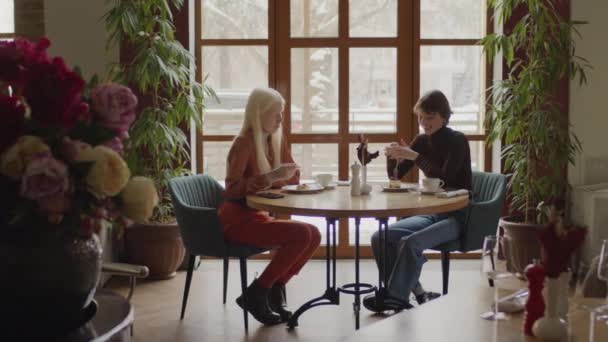 Largo tiro de dos diversas mujeres jóvenes sentadas a la mesa en un restaurante de lujo comiendo postres y charlando - Imágenes, Vídeo