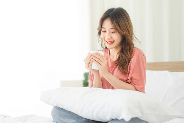 Serenidad de una joven asiática sosteniendo una taza de café. Disfrute de la tranquila mañana mientras se sienta en una cama blanca, con una sonrisa feliz. Momento de relajación y satisfacción. - Foto, imagen