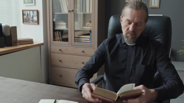Moderno sacerdote caucasico maturo seduto alla scrivania nel suo ufficio a leggere libri e scrivere citazioni nel suo quaderno - Filmati, video