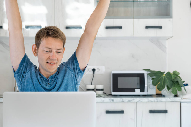 Wesoły młody przedsiębiorca pracujący na laptopie czuje satysfakcję z dobrej oferty i podnosi ręce. Freelancer inwestor zadowolony z dobrej inwestycji w domu biura. - Zdjęcie, obraz