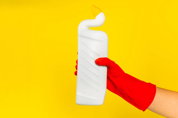 De hand van een schoonmaker in een rode rubberen handschoen die een fles met een reinigingsmiddel op een gele achtergrond vasthoudt. Commercieel schoonmaakbedrijf. Voorjaarsschoonmaak. Ruimte voor tekst of logo. - Foto, afbeelding