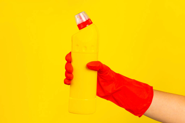 Το χέρι ενός καθαριστή σε ένα κόκκινο ελαστικό προστατευτικό γάντι κρατώντας ένα μπουκάλι με μια χημική ουσία καθαρισμού σε κίτρινο φόντο. Εμπορική εταιρεία καθαρισμού. Τακτικό καθάρισμα. Χώρος για κείμενο ή λογότυπο. - Φωτογραφία, εικόνα
