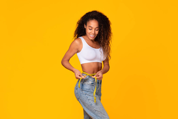Αφρο-Αμερικανίδα που δείχνει τη λεπτή κοιλιά της μετά από αδυνάτισμα, κρατώντας μεζούρα γύρω από τη μέση της πάνω από κίτρινο φόντο στούντιο. Υγιεινή διατροφή και προπόνηση αποτελέσματα ρουτίνας - Φωτογραφία, εικόνα
