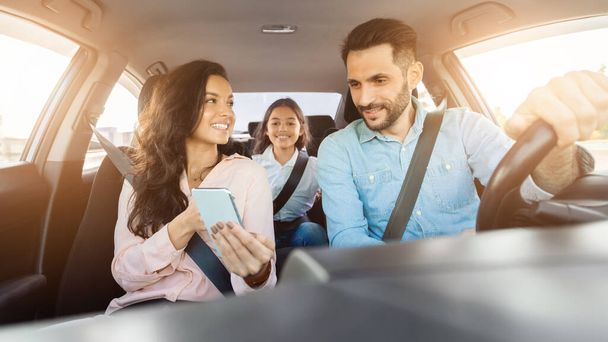 Wesoła rodzina cieszy się jazdy samochodem, matka trzyma smartfona, ojciec jazdy i ich córka siedzi z tyłu z zadowolonym wyrazem twarzy - Zdjęcie, obraz