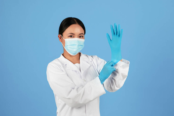 Mujer doctora asiática en máscara médica poniéndose guantes médicos y mirando a la cámara, confiada joven doctora en bata de laboratorio posando aislada sobre fondo azul del estudio, espacio para copiar - Foto, Imagen