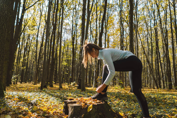 The girl ties her shoelaces. Beautiful girl doing fitness in nature on a sunny autumn forest. Body positive, sports for women, harmony, healthy lifestyle, self-love and wellness. - Photo, Image