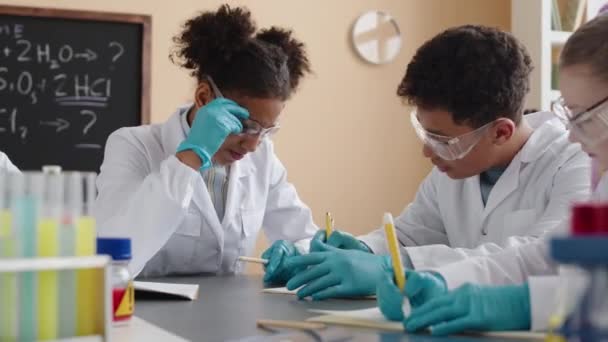 Vita di scolari multirazziali in camici e occhiali di sicurezza che chattano durante l'esperimento di laboratorio chimico nella moderna classe di scienze - Filmati, video