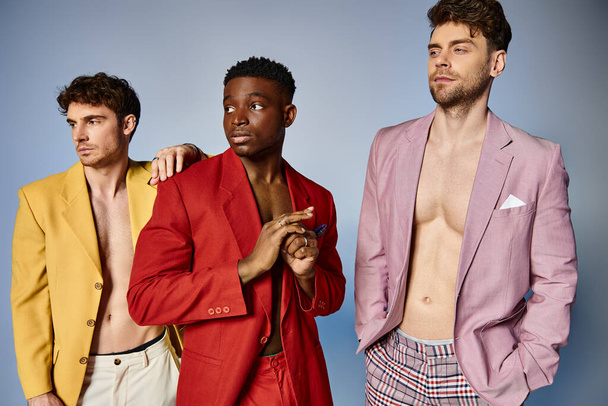 three multicultural men in vibrant unbuttoned suits posing together on gray backdrop, fashion - Photo, Image