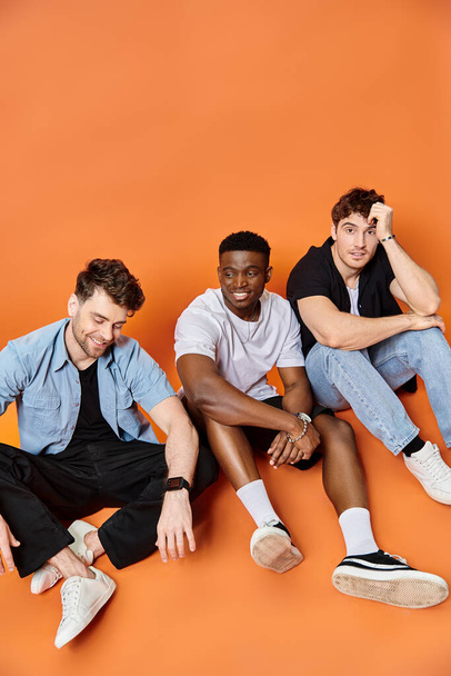 three joyous multiracial men in urban casual clothes smiling and sitting on floor, fashion concept - Photo, Image