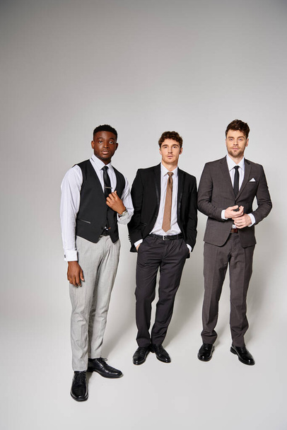 красивые молодые мультикультурные друзья в умных деловых костюмах позируют вместе на сером фоне - Фото, изображение