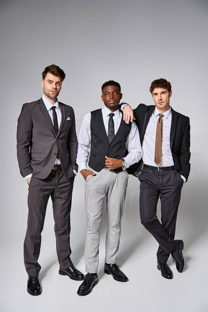gut aussehende junge diverse Freunde in schicken Business-Anzügen posieren gemeinsam vor grauem Hintergrund - Foto, Bild