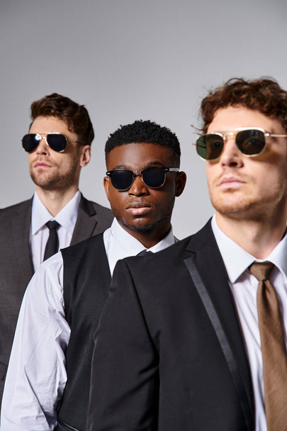 handsome multiracial male models in elegant smart suits with sunglasses posing on gray backdrop - Photo, Image