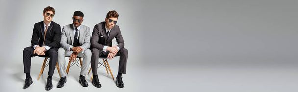 atrakcyjne różnych mężczyzn w okularach przeciwsłonecznych w odzieży biznesowej siedzi na krzesłach na szarym tle, sztandar - Zdjęcie, obraz
