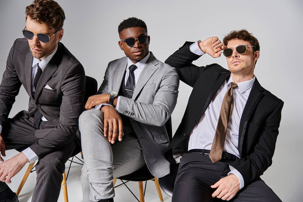 bellissimi uomini interrazziali con occhiali da sole in abiti eleganti seduti su sedie su sfondo grigio - Foto, immagini