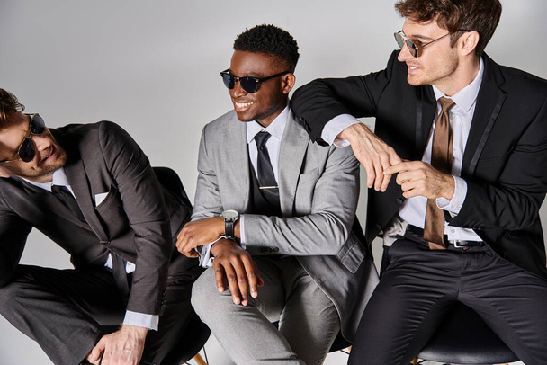 šťastný pohledný multikulturní muži v podnikání chytré obleky sedí na židlích a radostně se usmívá - Fotografie, Obrázek