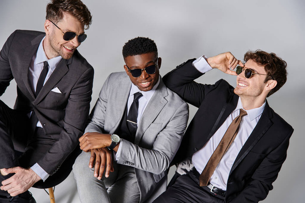 fröhliche attraktive multikulturelle Männer in schicken Business-Anzügen sitzen auf Stühlen und lächeln fröhlich - Foto, Bild