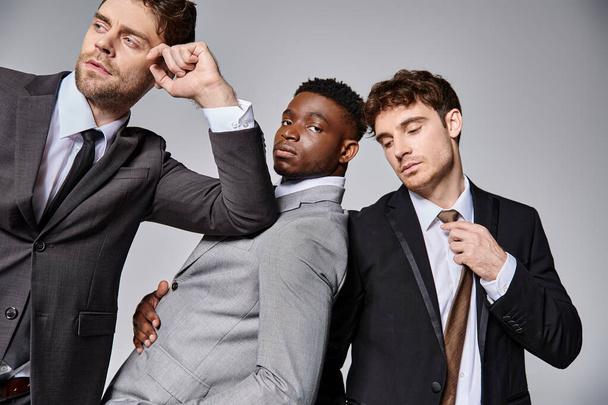jóvenes hombres multiculturales de buen aspecto en trajes inteligentes de negocios posando juntos sobre fondo gris - Foto, imagen