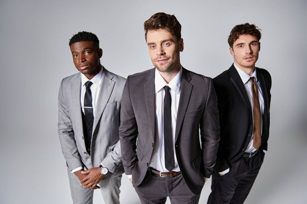 atractivo jóvenes modelos masculinos multiculturales en trajes casuales de negocios posando sobre fondo gris - Foto, imagen
