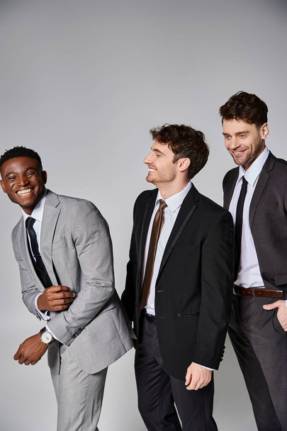 goed uitziende vrolijke multiculturele mannelijke modellen in slimme pakken glimlachen oprecht op grijze achtergrond - Foto, afbeelding