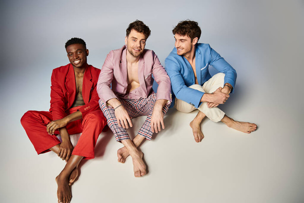 alegres amigos interracial en vibrantes trajes de colores sentados en el suelo con las piernas cruzadas, la moda - Foto, Imagen