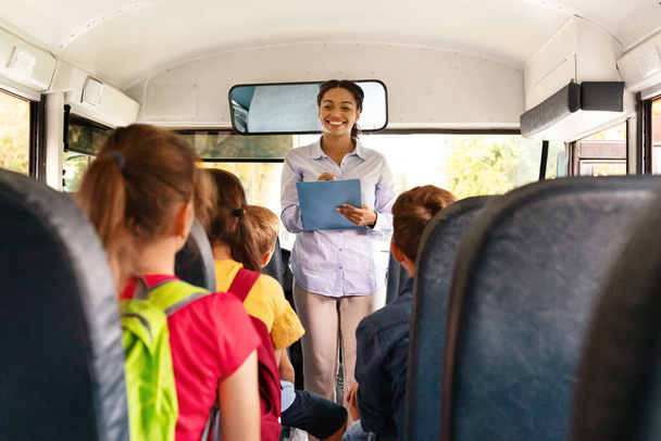 Vriendelijke zwarte lerares die de namen van de kinderen afvinkt in de schoolbus, groep kinderen die op hun stoel zitten in het voertuig, wachtend op de reis, klaar om samen te reizen, vrije ruimte - Foto, afbeelding