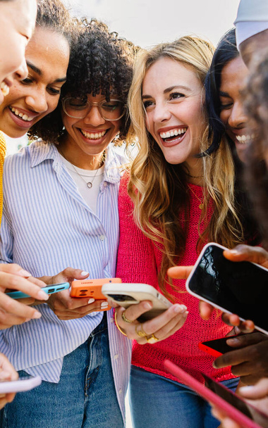 Χαρούμενη γυναικεία ομάδα που χρησιμοποιεί το κινητό τηλέφωνο μαζί σε εξωτερικούς χώρους. Διαφορετικές φίλες γελάνε ενώ βλέπουν αστείο περιεχόμενο στα μέσα κοινωνικής δικτύωσης σε εφαρμογή δικτύου κινητής τηλεφωνίας. Έννοια της φιλίας και της τεχνολογίας - Φωτογραφία, εικόνα