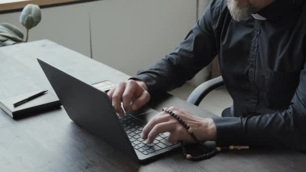 Wysoki kąt ujęcia dojrzałego kaukaskiego księdza noszącego okulary i słuchawki bezprzewodowe, trzymającego różaniec, siedzącego przy biurku w swoim biurze piszącego coś na laptopie podczas rozmowy telefonicznej - Materiał filmowy, wideo